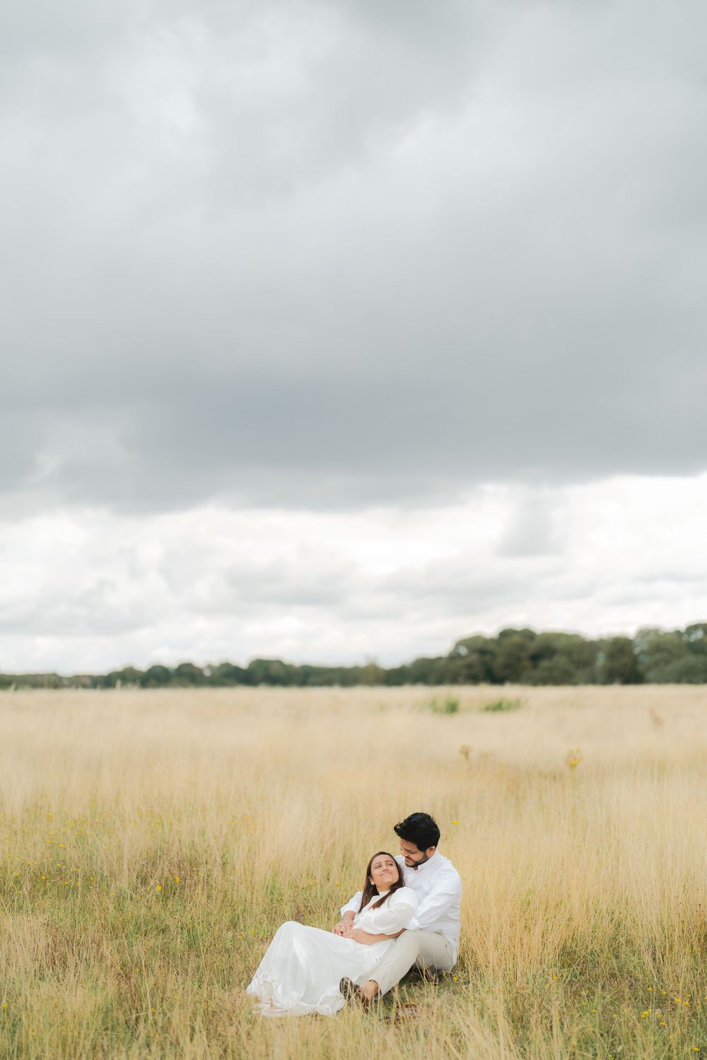 Couple in white sitting together in a golden meadow under dramatic cloudy skies.
