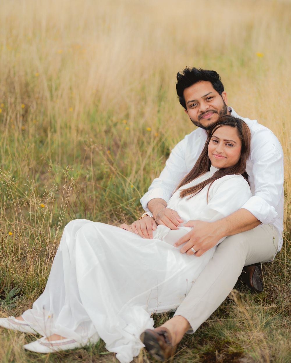 A couple in matching white outfits sitting in a golden grass field, woman leaning against man, smiling warmly.