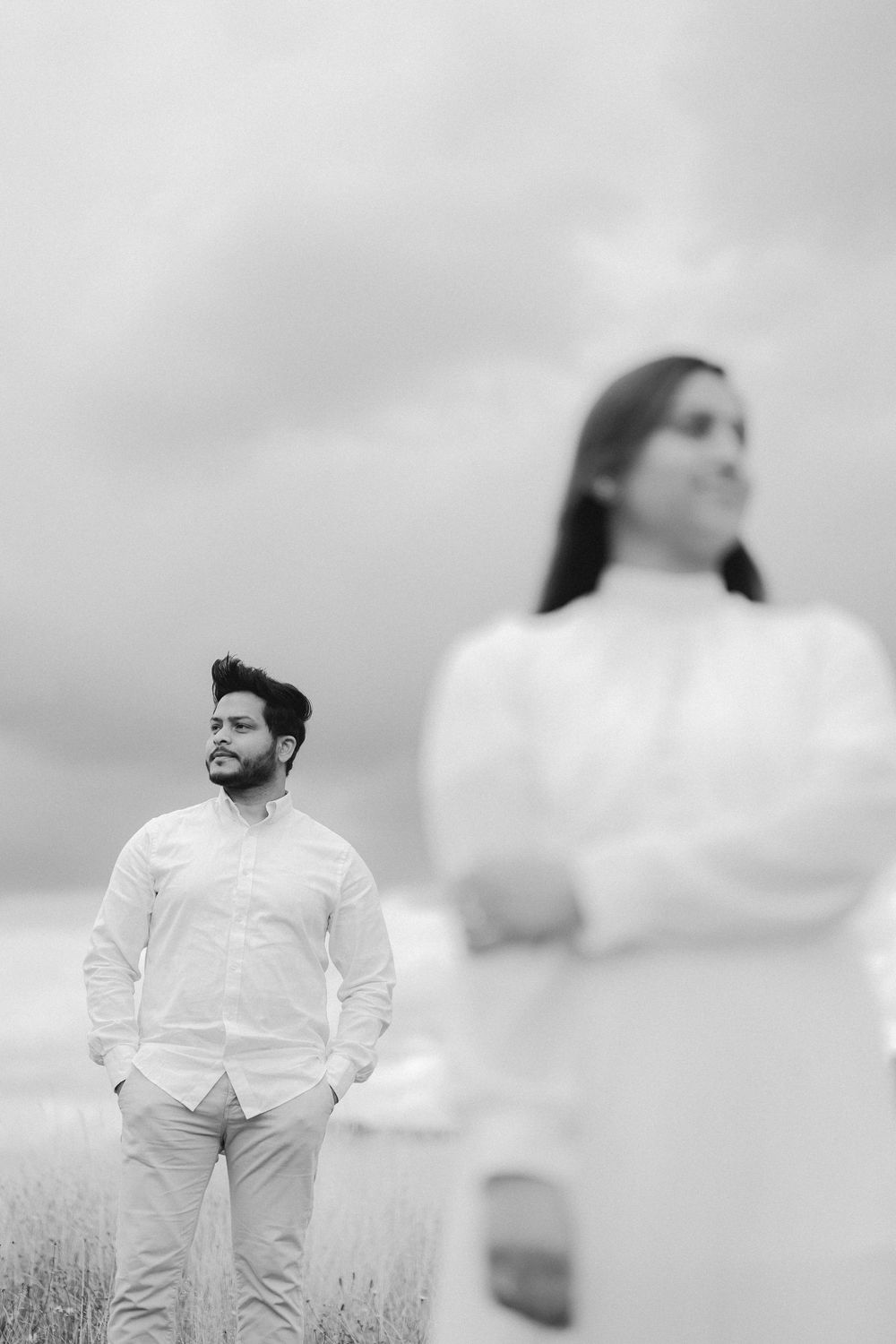 Black and white photo of a bearded man in white shirt standing in field, woman with crossed arms blurred in foreground.
