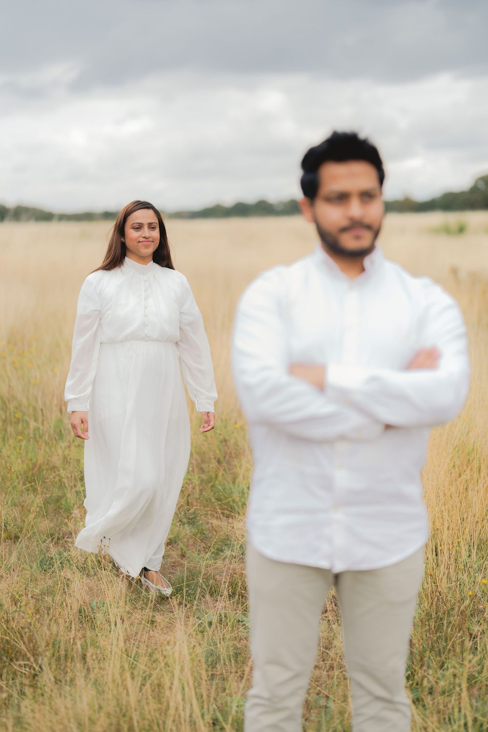 A woman in a white dress walks toward a man in a white shirt standing in a golden field under cloudy skies.