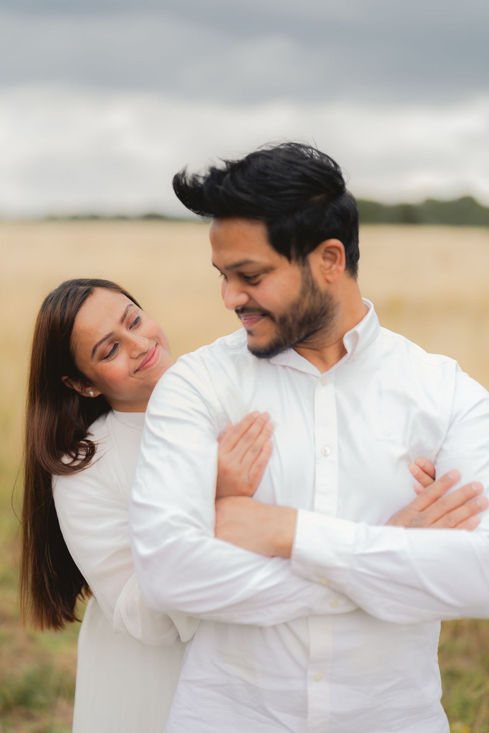 A couple in matching white outfits embraces in a golden field, the woman resting her head on the man's shoulder.