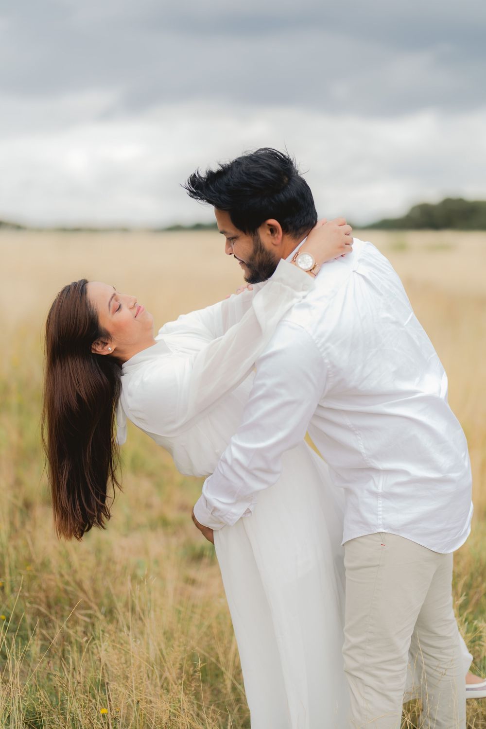 A couple in white outfits embrace romantically in a golden field, the man lifting the woman as she leans back joyfully.