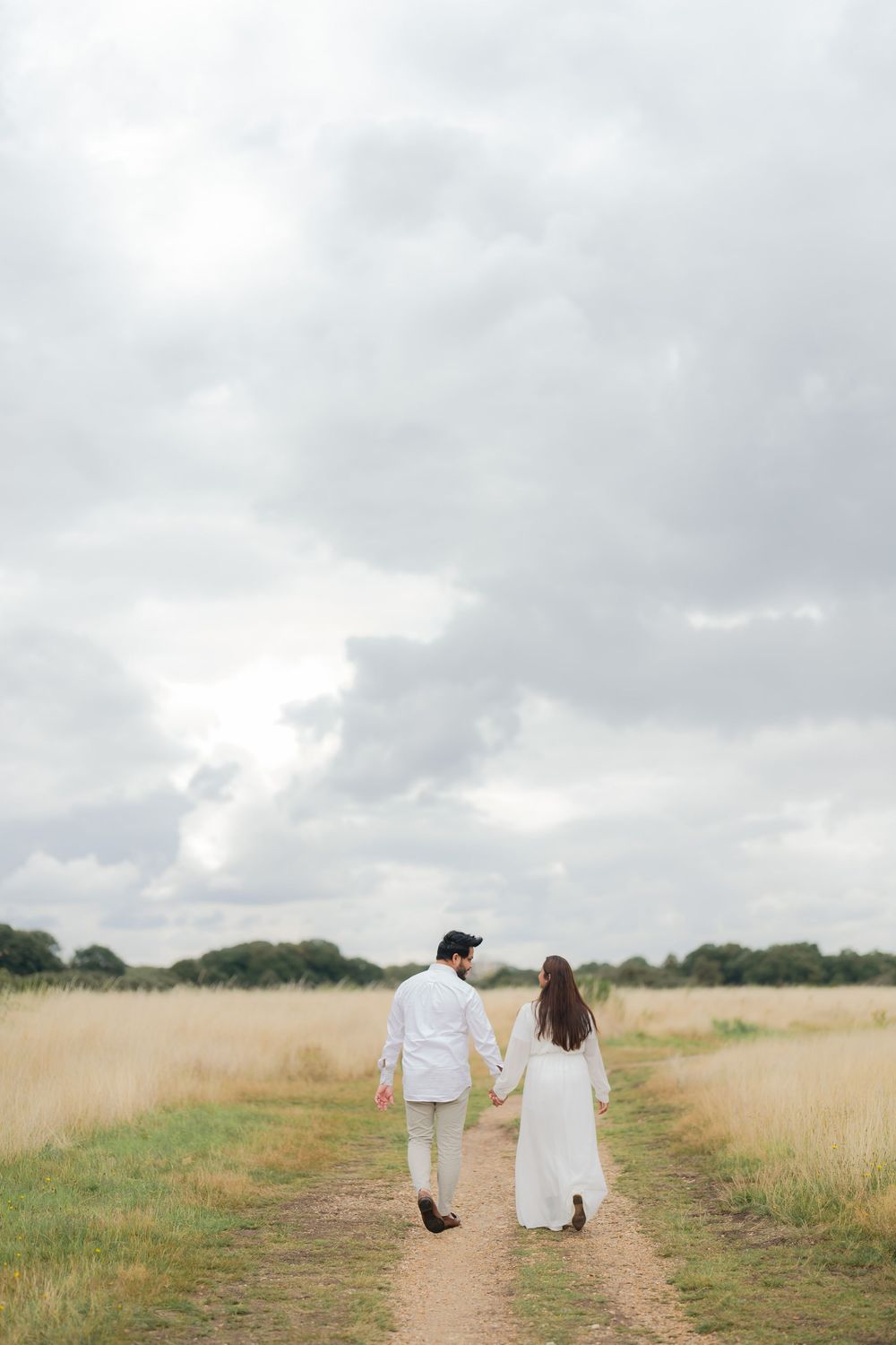 Couple in white holding hands walking down a dirt path through golden fields under a cloudy sky.