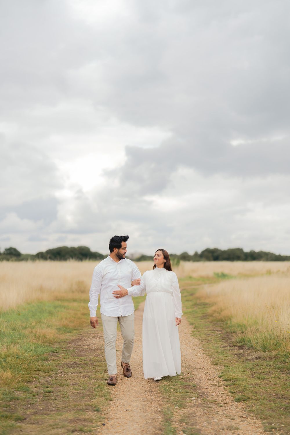 Couple in white outfits walking hand in hand on a dirt path through golden fields under a cloudy sky.