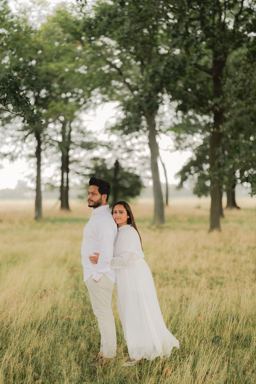 Couple in matching white outfits embracing in a lush green field with tall trees in the background.