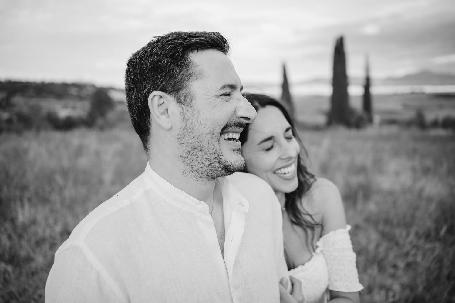 Black and white photo of a laughing couple embracing in a field with cypress trees in the background in Elopement in Val D'Orcia