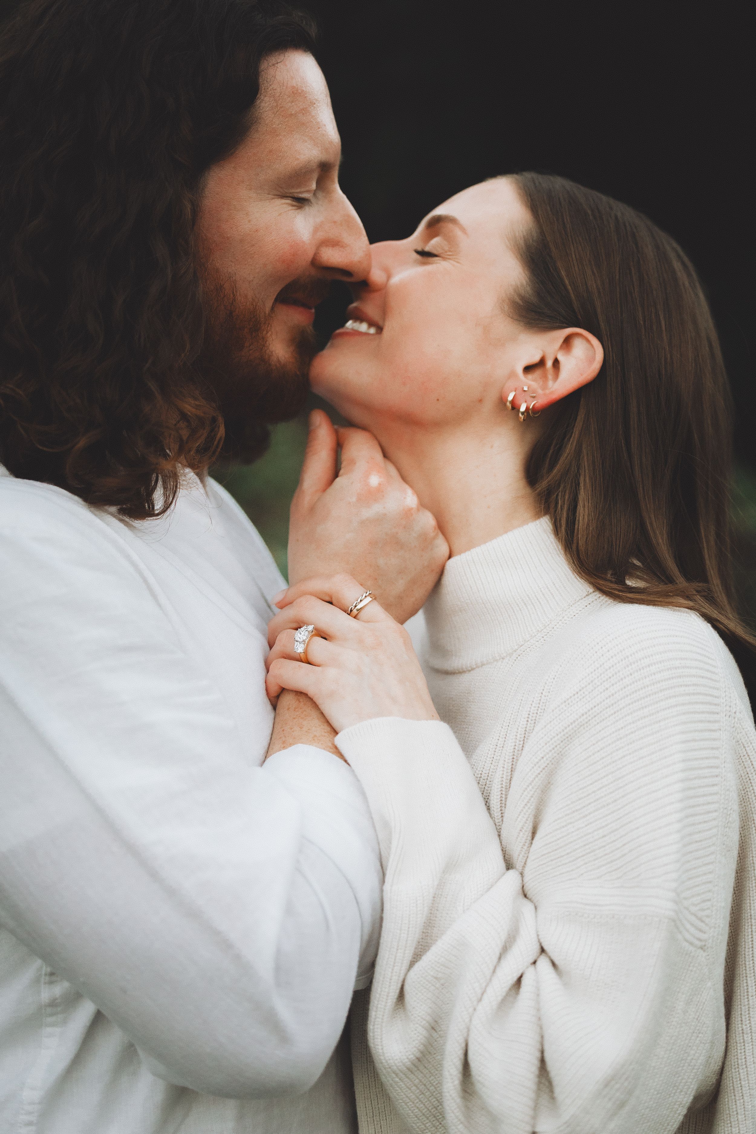 Romantic couple in white sweaters sharing a tender moment, woman wearing an engagement ring, eyes closed, nearly kissing.
