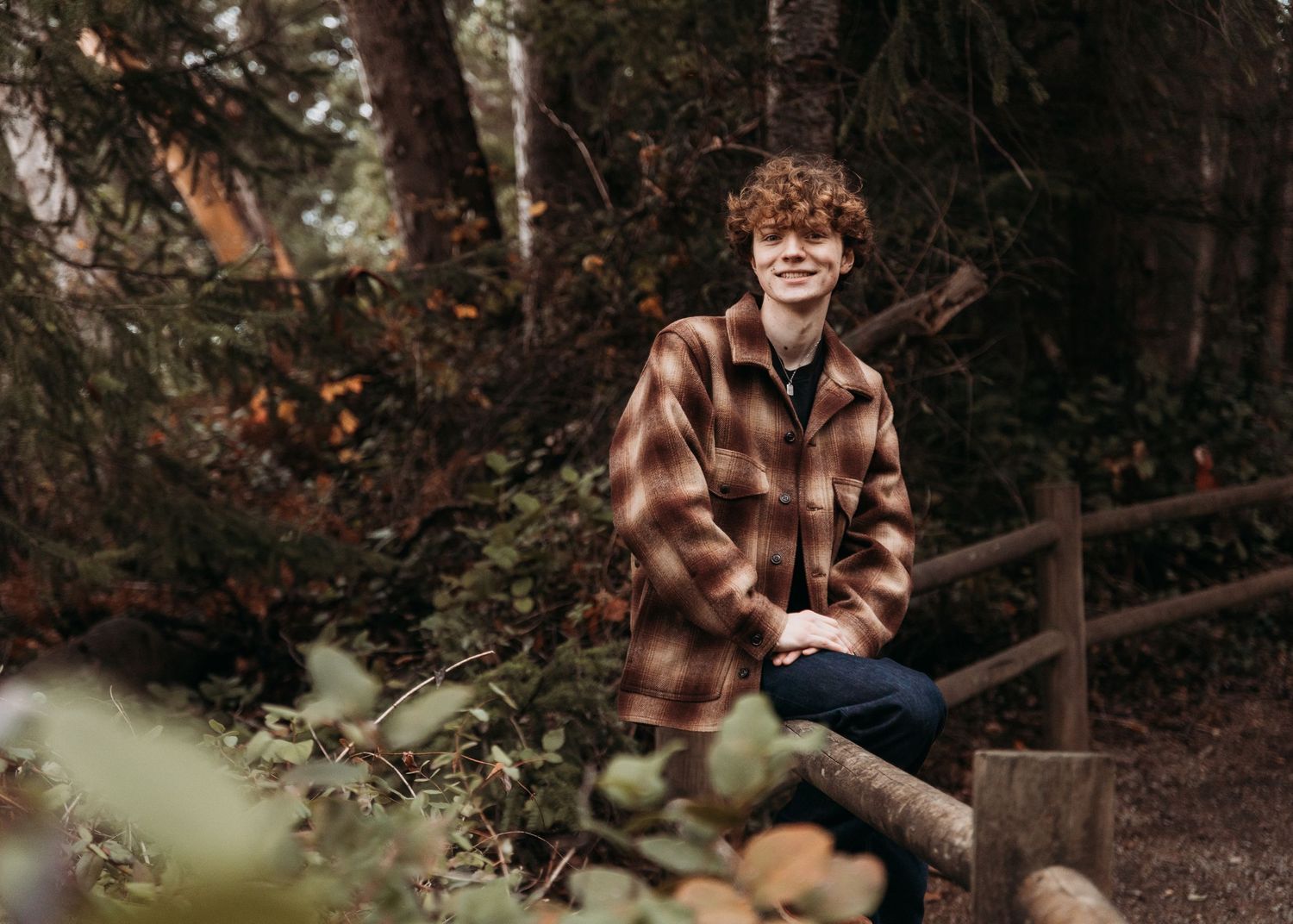 Young man in plaid flannel jacket smiling outdoors on a wooden fence surrounded by lush forest greenery.