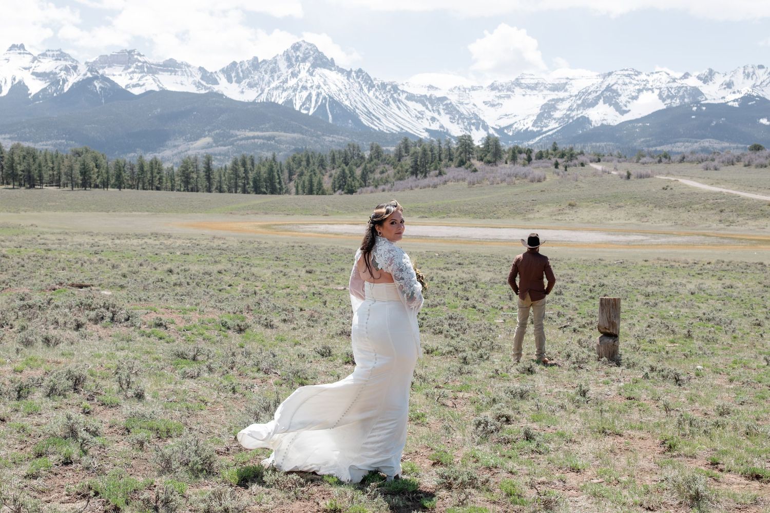 Bride in white gown with floral crown stands in mountain meadow with snow-capped peaks and groom in background. She looks back at the camera
