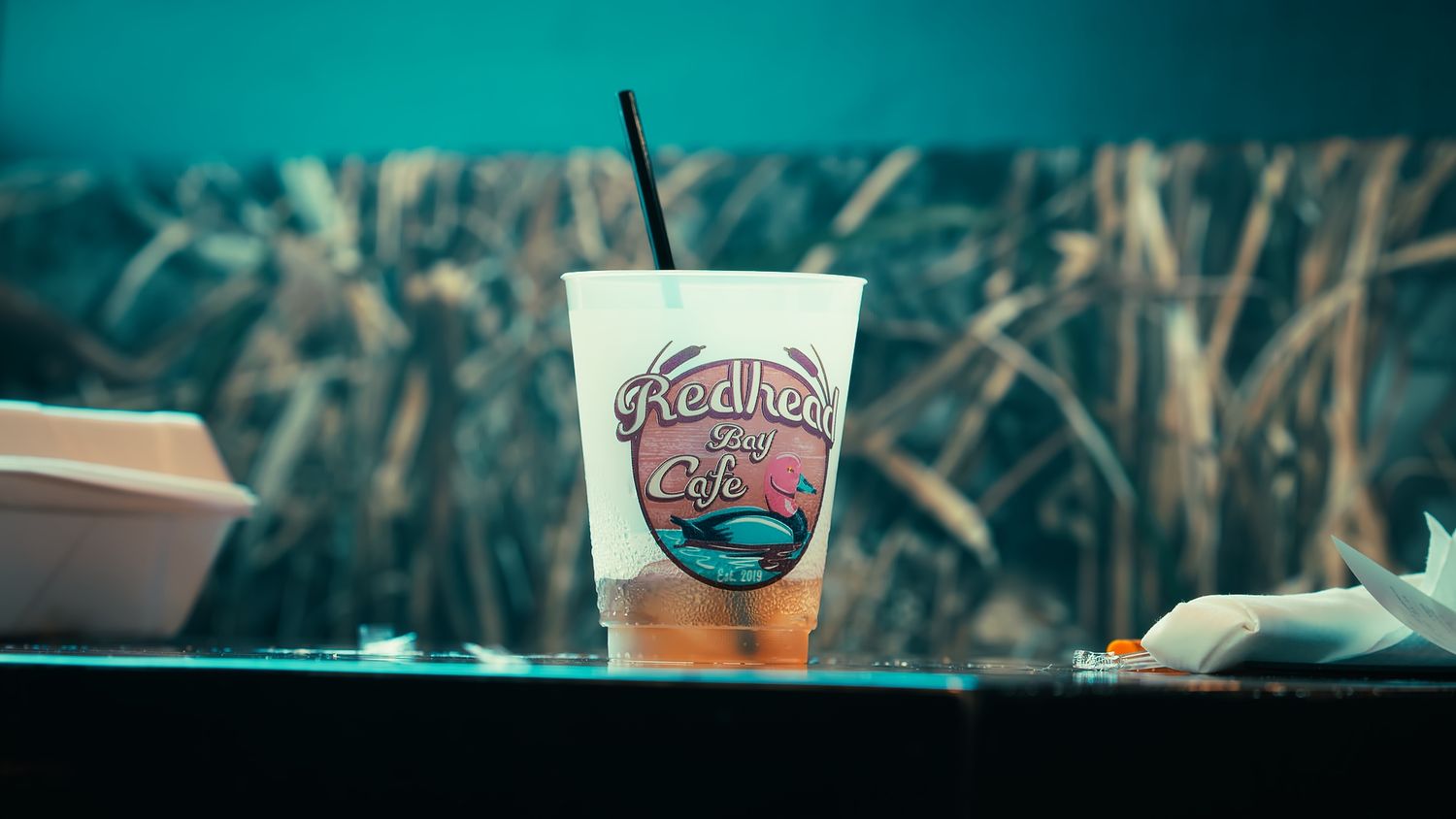 Redhead Bay Cafe restaurant branded cup with a straw sitting on a table outdoors.