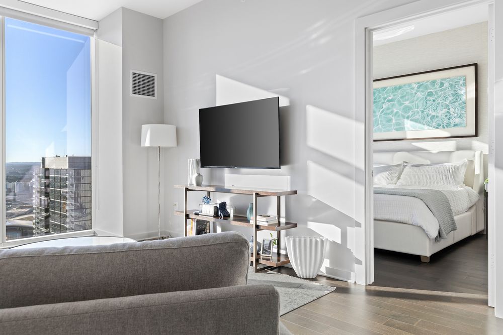 Modern apartment living room with city views, wall-mounted TV, and connecting bedroom visible through doorway.