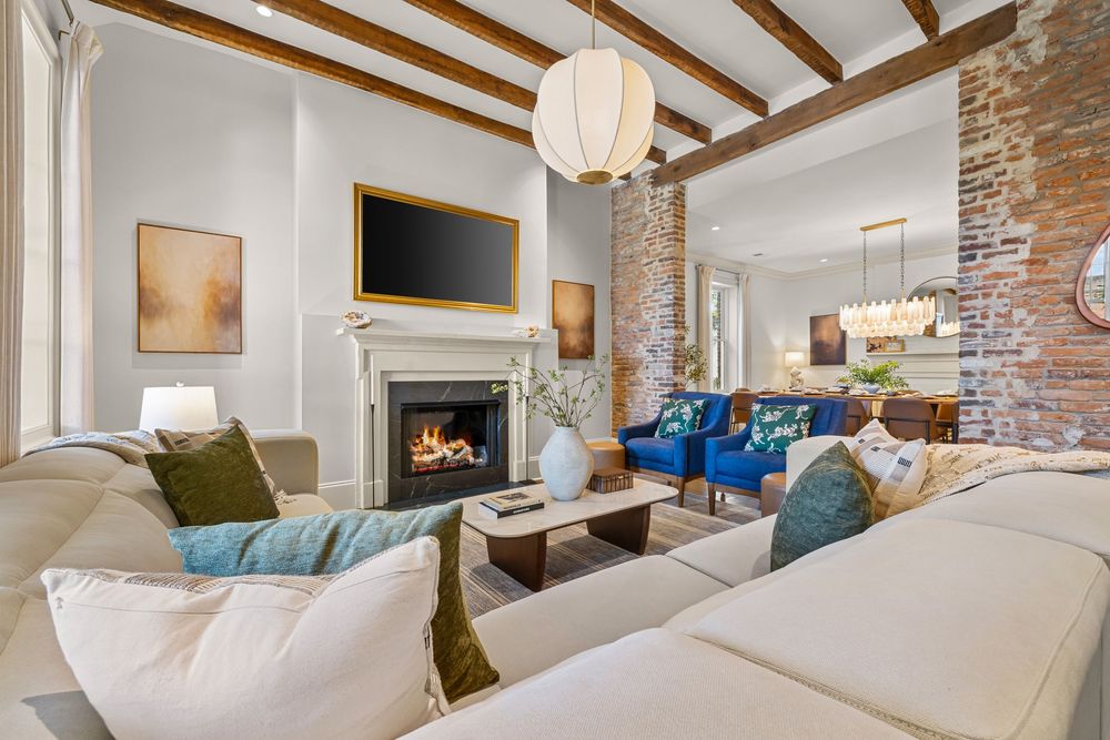Elegant living room with exposed beam ceiling, lit fireplace, blue velvet sofa, white sectional, and rustic brick accent wall.