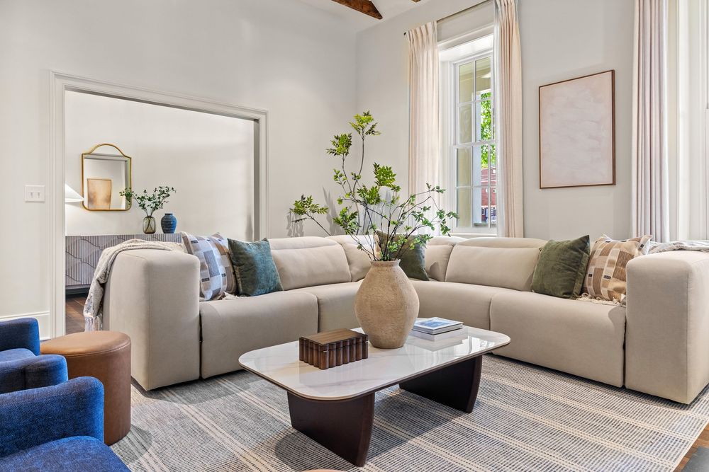 Modern living room with beige sectional sofa, marble coffee table, blue accent chairs, and natural light from tall windows.