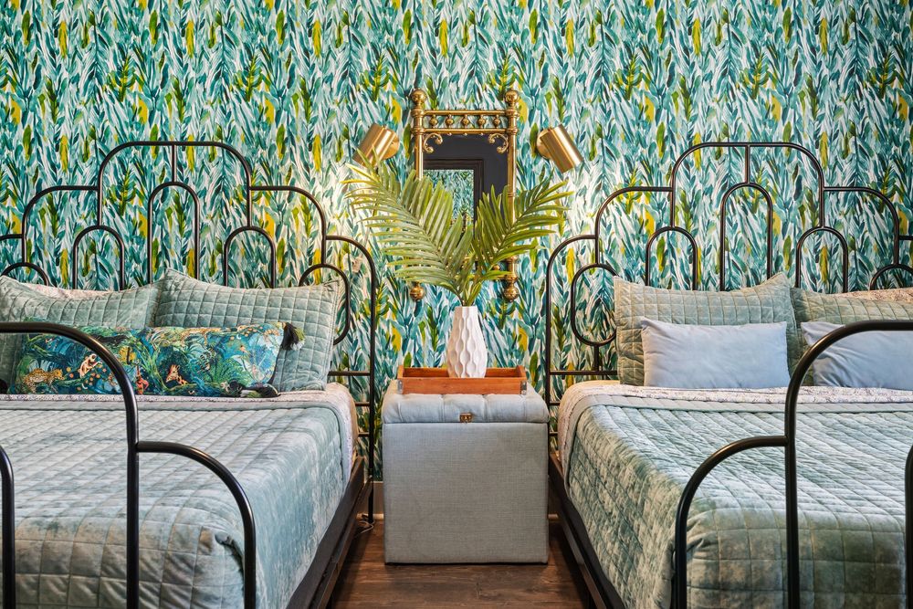 Two twin beds with green quilted bedding flank a nightstand against vibrant tropical leaf wallpaper in a stylish bedroom.