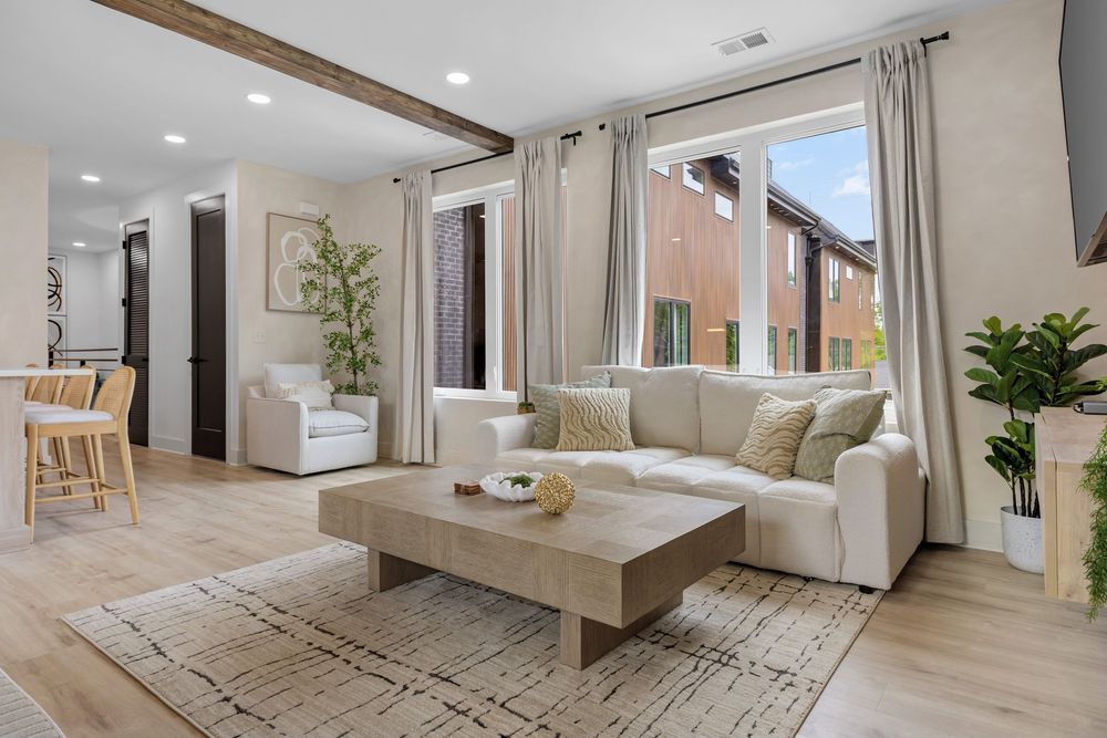 Bright modern living room with white sofa, wooden coffee table, exposed beams, and large windows with sheer curtains.