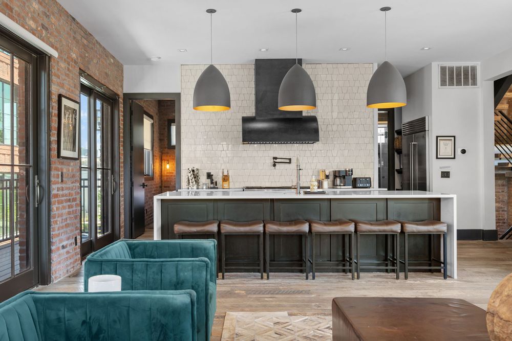 Modern open-concept kitchen with gray pendant lights, dark island, bar stools, brick walls, and teal velvet sofa.
