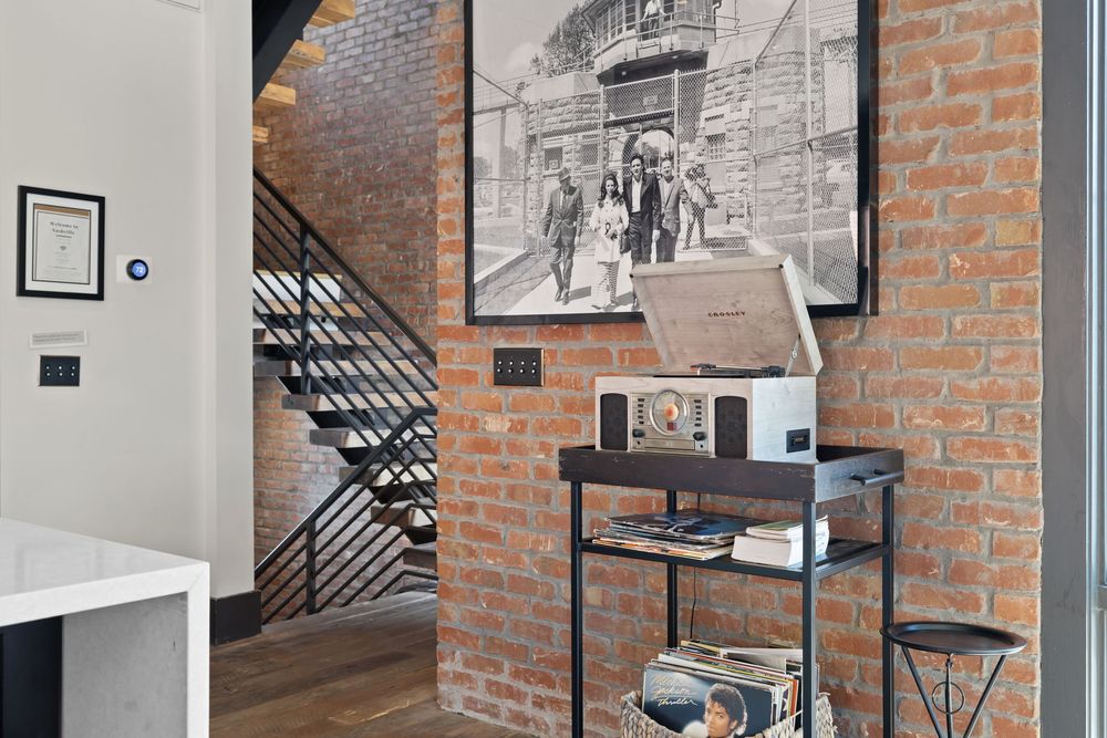 Industrial loft entryway with exposed brick wall, black metal shelving unit, vintage B&W photo art, and modern staircase.