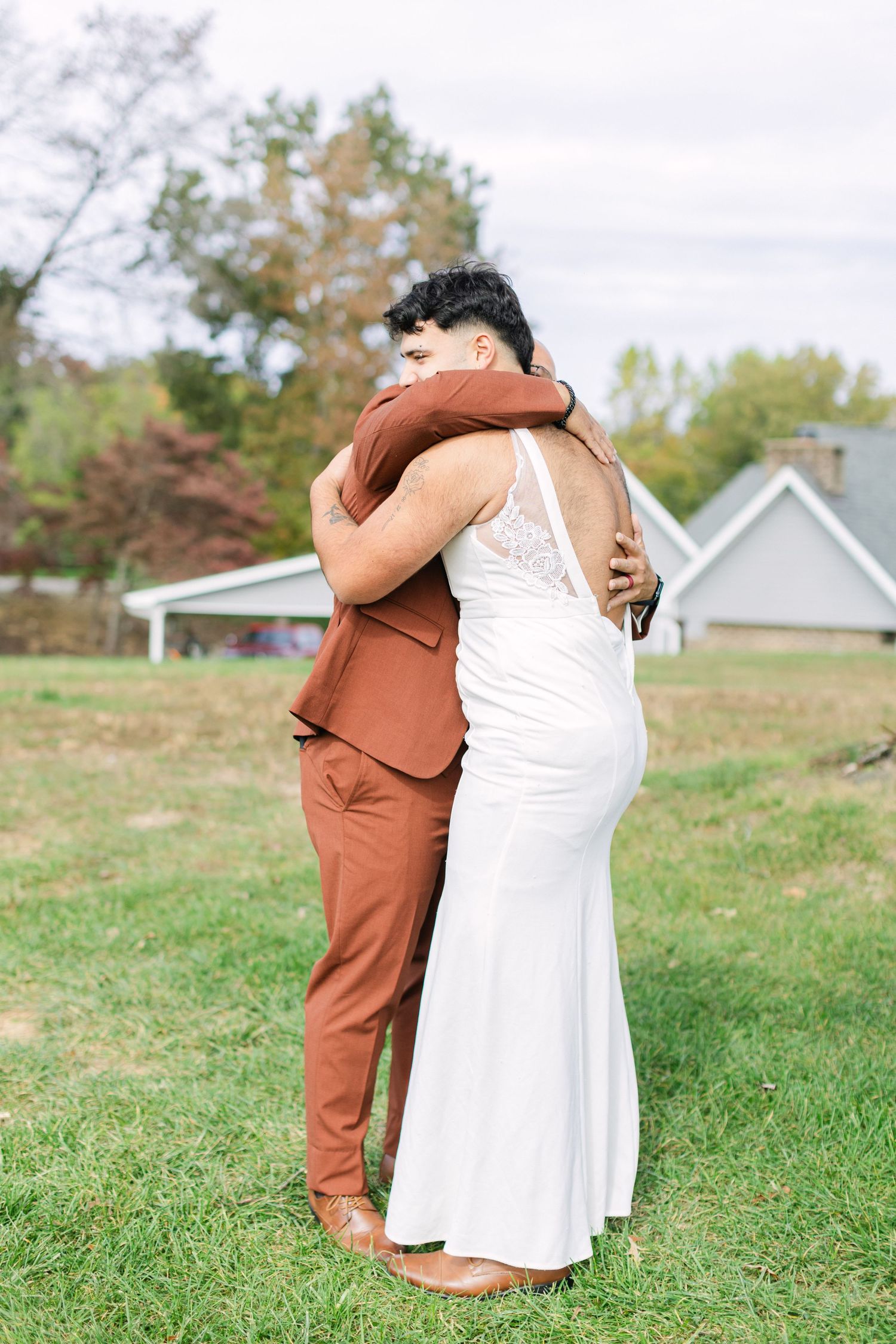 Bride in white gown and groom in rust suit share a heartfelt embrace outdoors on green grass.