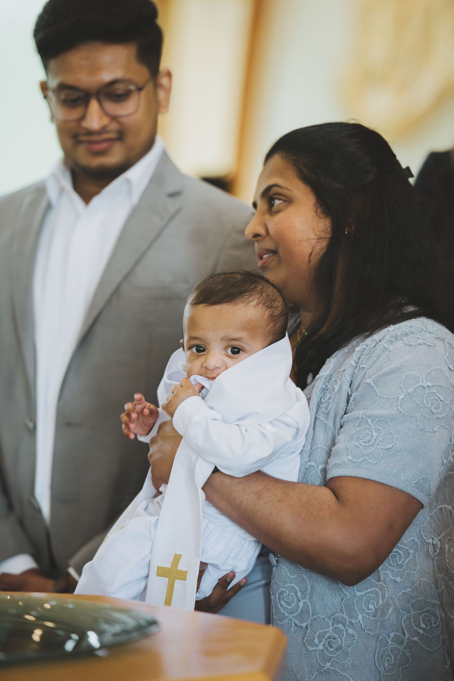 Family at a baby baptism ceremony, mother holding infant in white christening gown with gold cross in a church.