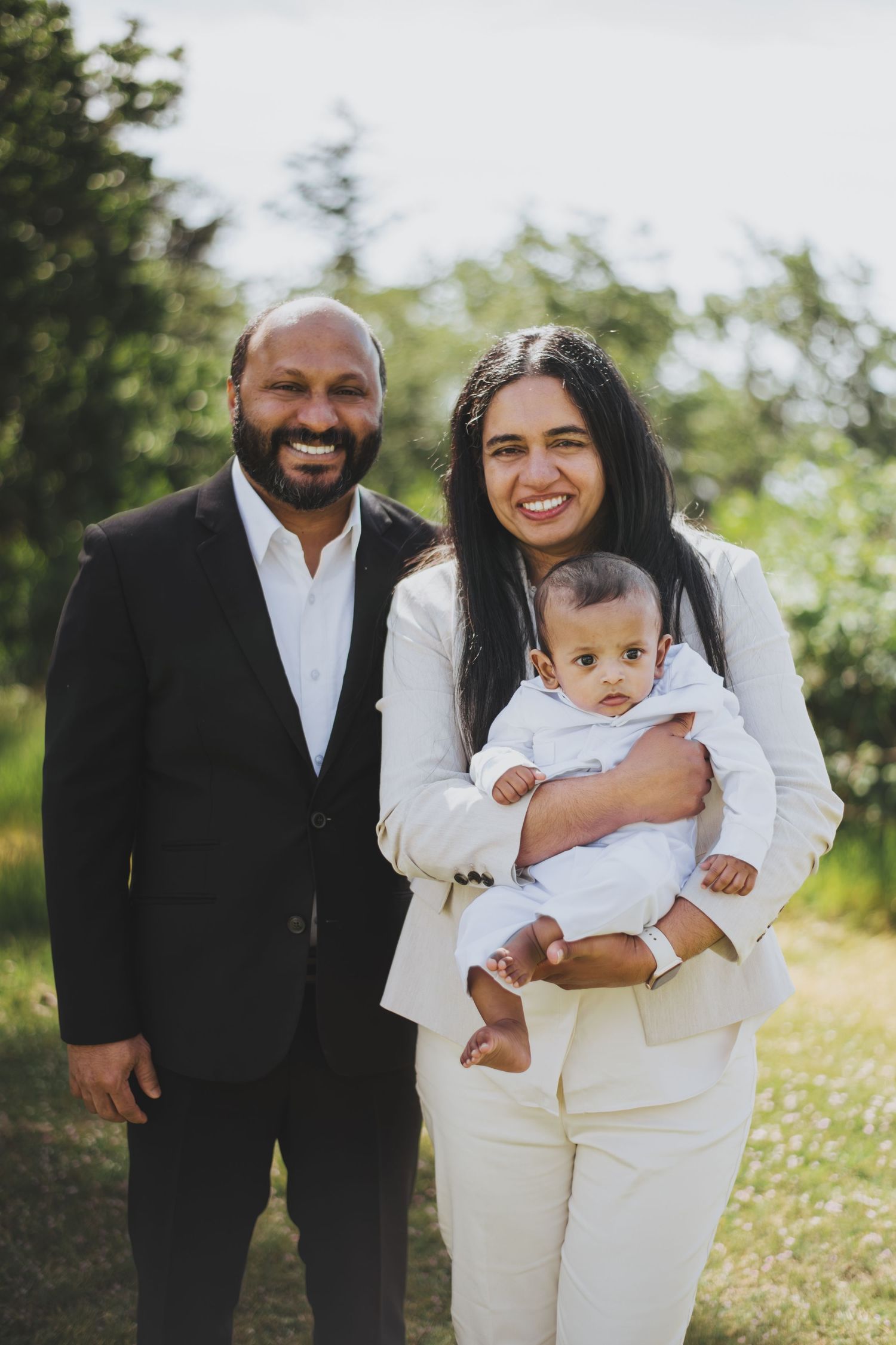 Smiling South Asian family portrait outdoors, parents in formal attire holding a baby dressed in white.