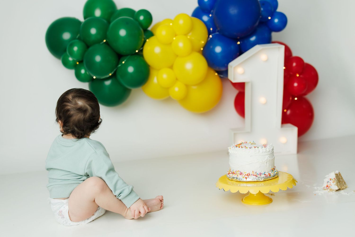 Baby sits before a lit number 1, white birthday cake, and colorful balloon garland for a first birthday photo.