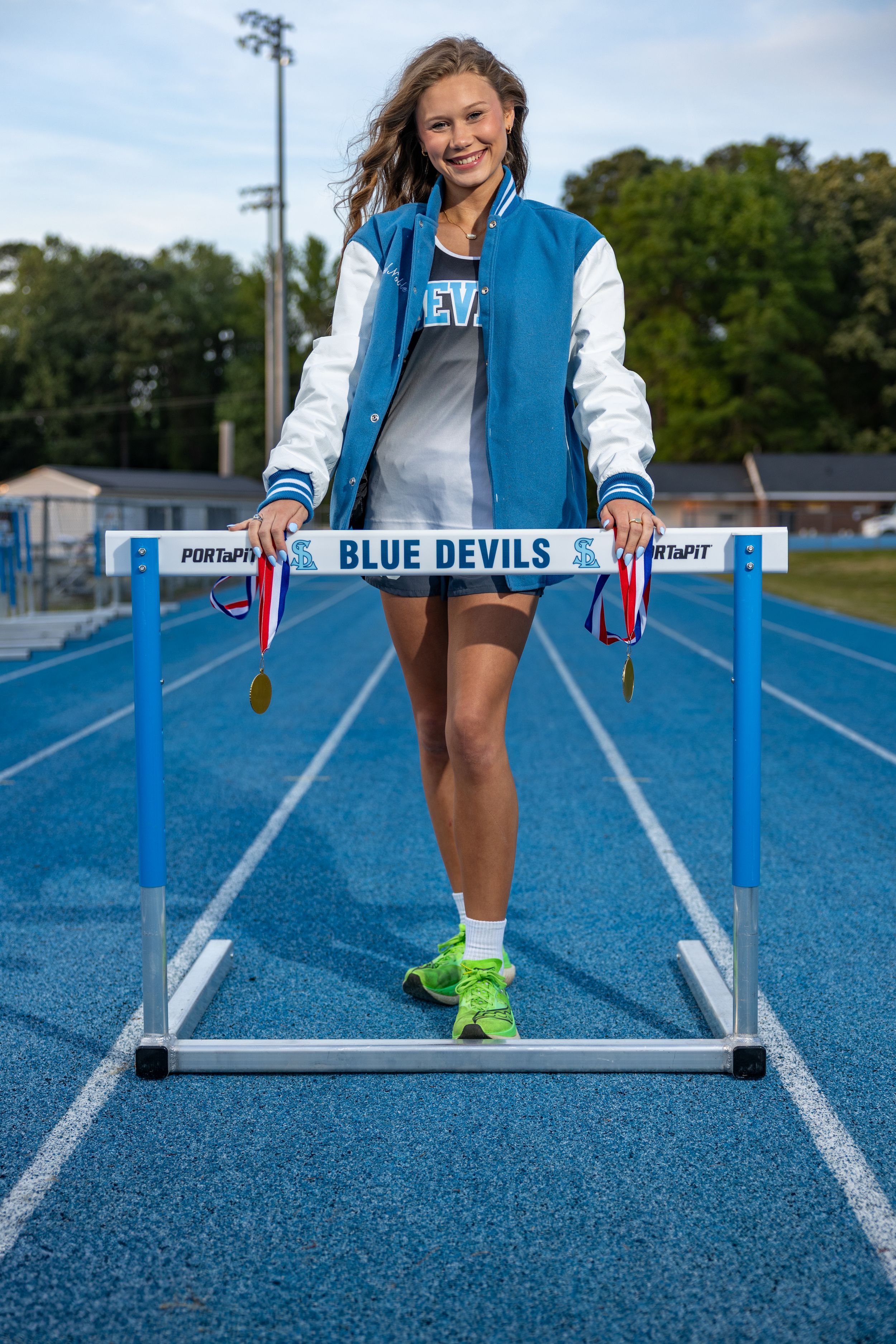 Young female athlete poses with Blue Devils track hurdle, wearing varsity jacket and medals on a blue running track.