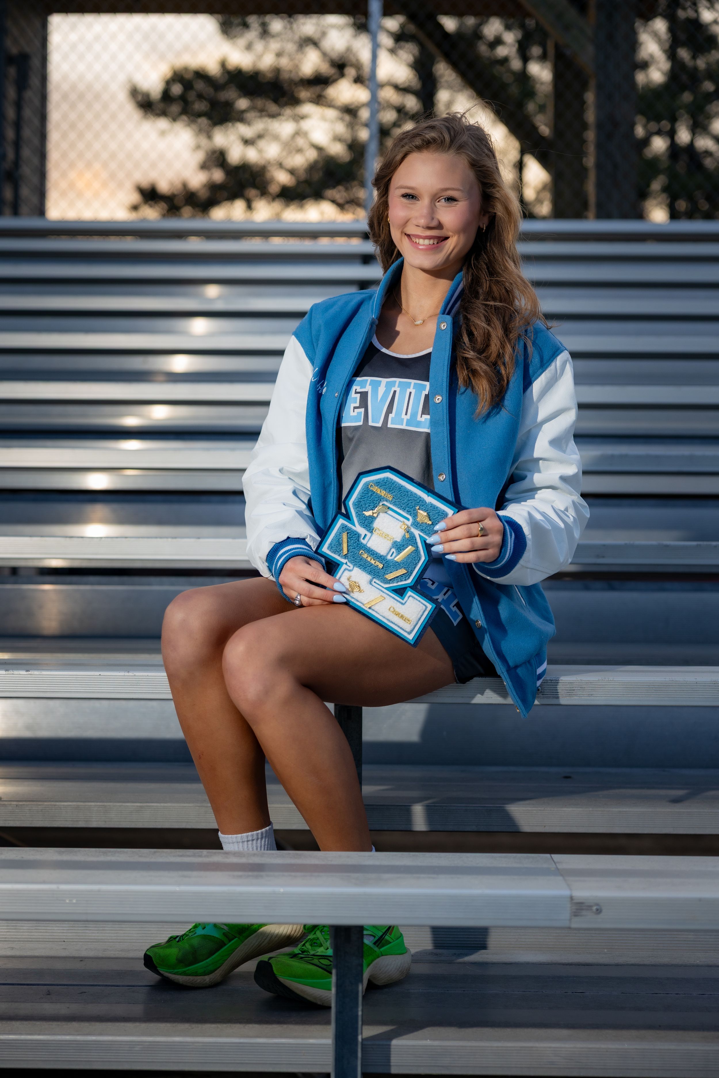 Smiling female athlete in blue Devils letterman jacket sits on bleachers holding a varsity letter award, wearing green sneakers.