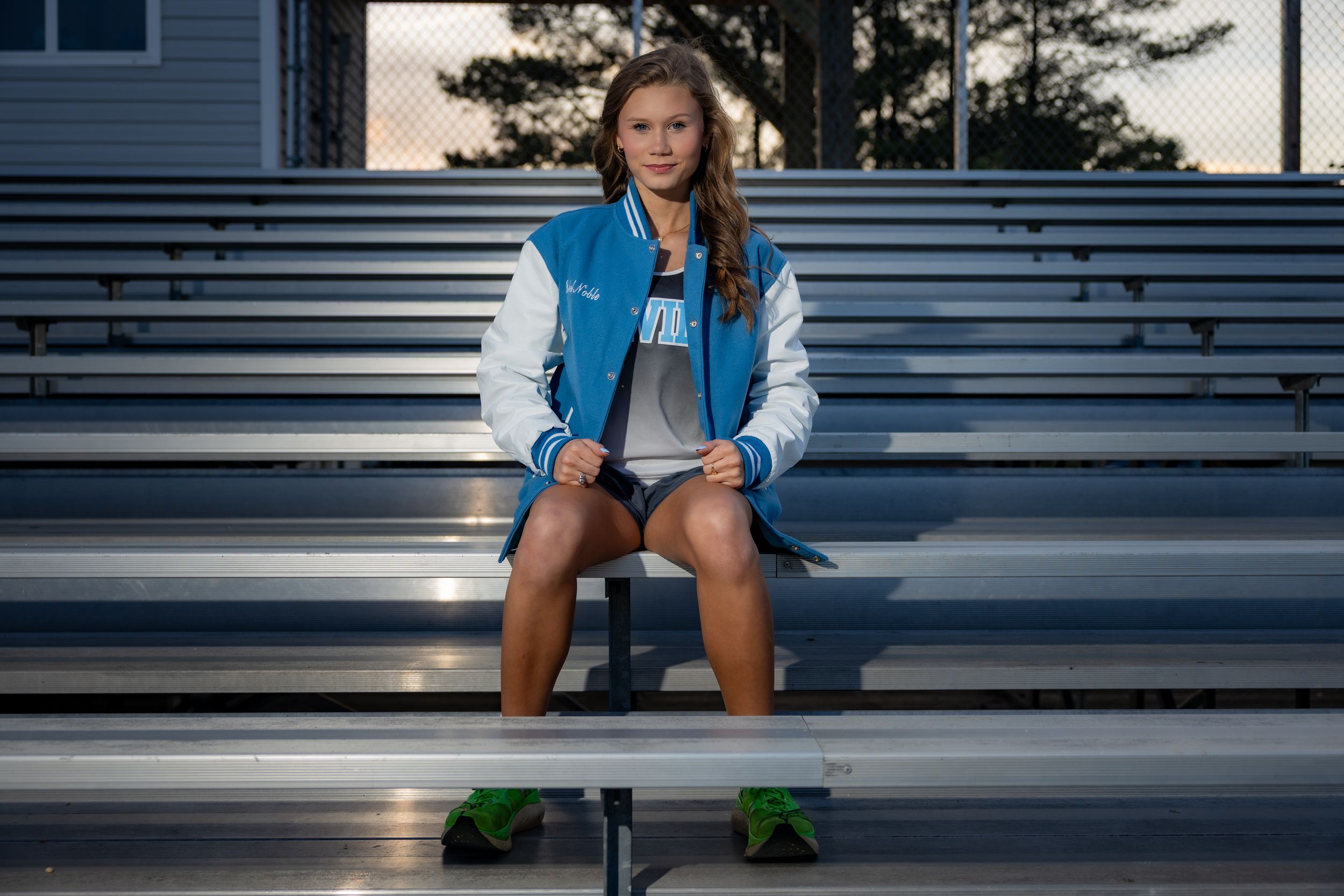 Young woman in blue varsity jacket sits on metal bleachers outdoors, posing for a sports portrait photo.