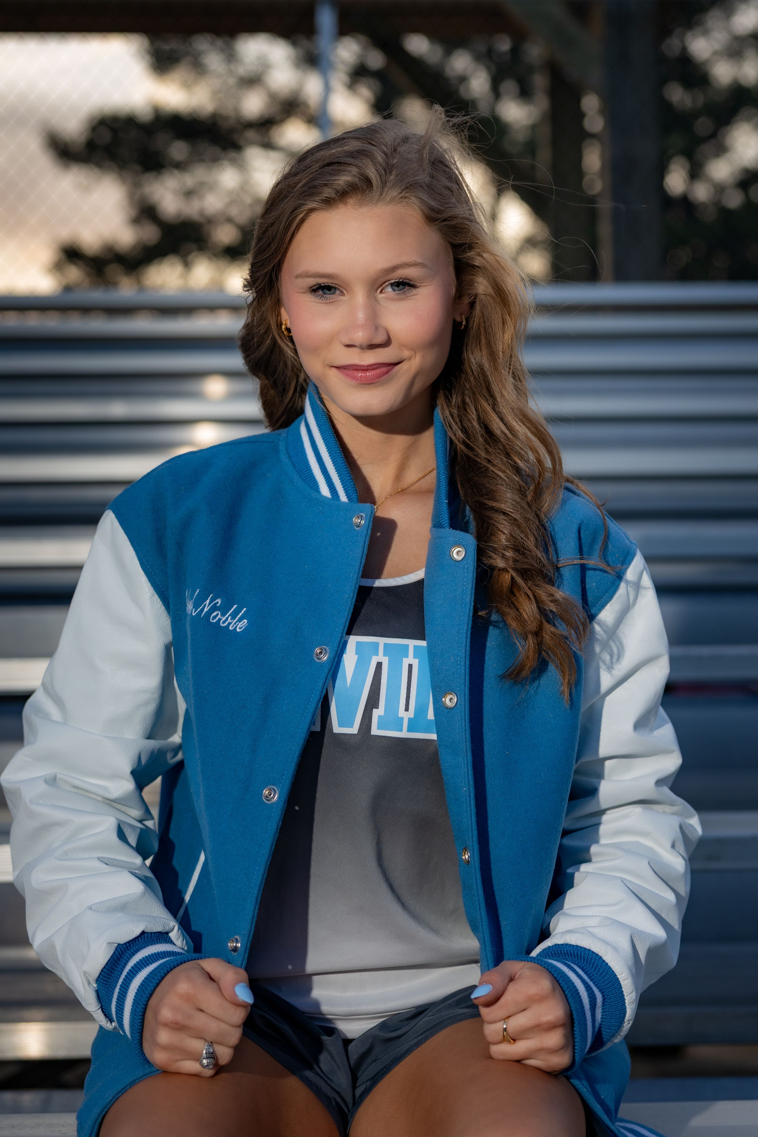 Young woman in blue and white varsity jacket sitting on bleachers, smiling confidently outdoors.