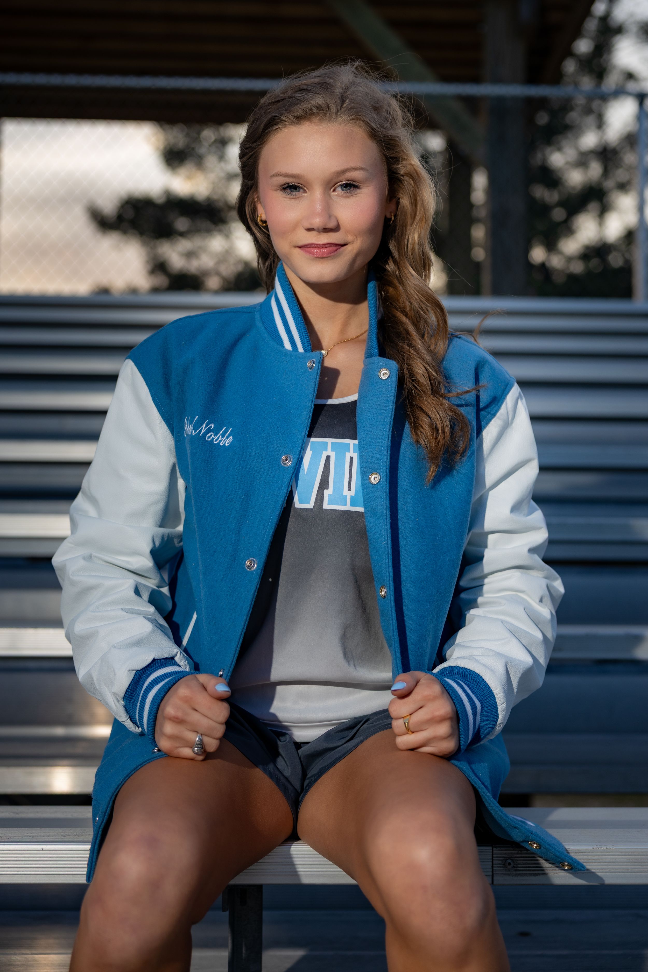 Young woman in blue and white varsity jacket sitting on bleachers, smiling confidently at camera.