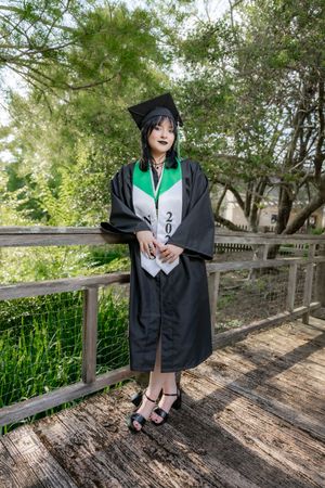 Graduate in black cap and gown with green stole posing on wooden bridge surrounded by lush green trees.