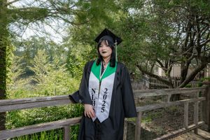 Graduate in black cap and gown with green stole and 2025 sash poses outdoors near wooden fence.