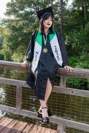 Graduate in black cap and gown with green sash and 2025 stole poses on wooden bridge outdoors.
