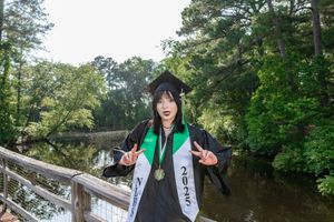 Graduate in cap and gown with 2025 sash poses on bridge surrounded by lush green trees near a river.