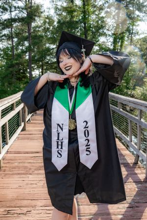 Student in black graduation gown with NLHS 2025 stole and medal poses on wooden bridge surrounded by trees.