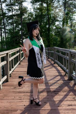 Young woman in black dress with NJHS sash and cat ears poses on wooden bridge surrounded by trees.