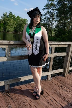 A young woman in a black dress, graduation cap, and 2025 stole poses on a wooden dock by a lake.