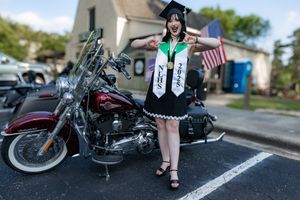 Graduate in cap and gown poses excitedly beside a Harley-Davidson motorcycle holding an American flag outdoors.