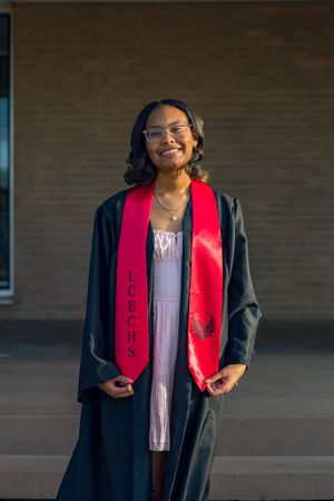 Smiling graduate in black gown and red LCECHS stole poses outdoors for graduation portrait photo.