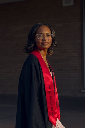 Young woman in black graduation gown and red stole with glasses smiling outdoors.