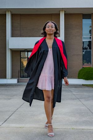 Young Black woman in black and red graduation gown walks confidently in pink dress and heels outside brick building.