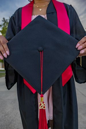 Graduate holding black mortarboard cap with red tassel and 26 charm in graduation gown.