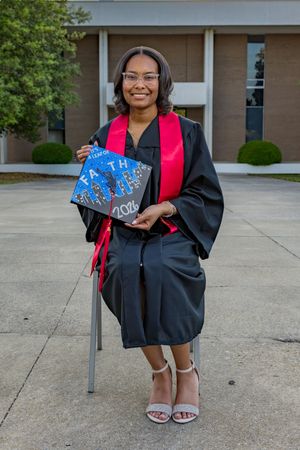 Smiling graduate in black robe with red stole holds decorated 2024 cap outside university building.