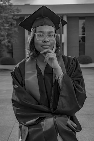 Young woman in graduation cap and gown poses thoughtfully outdoors in black and white portrait photo.