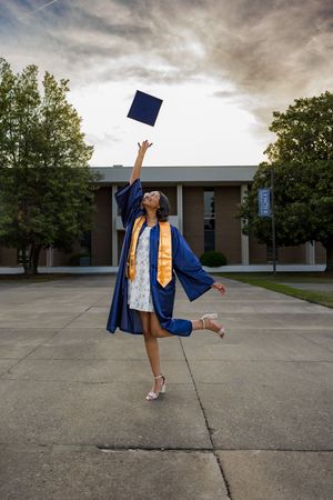Joyful graduate in blue cap and gown tossing mortarboard into cloudy sky outdoors.