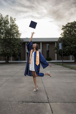 Joyful graduate in blue cap and gown tossing mortarboard into cloudy sky outdoors.