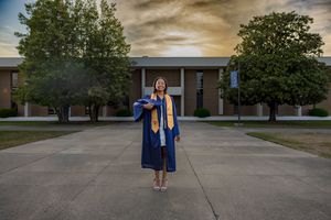 Graduate in blue cap and gown poses proudly on campus plaza at sunset with brick building backdrop.