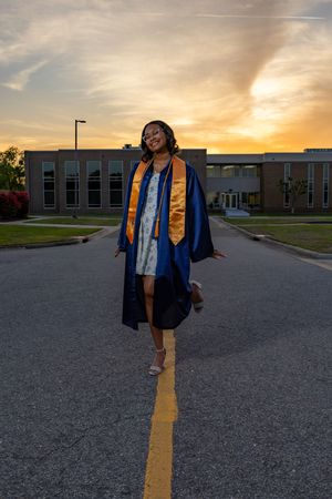 Smiling graduate in blue gown and gold stole walking on road at golden sunset.