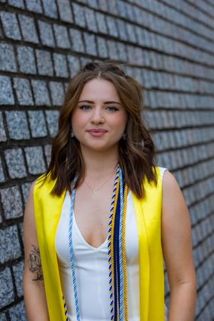 Young woman in yellow graduation stole and white dress smiling in front of a brick wall.