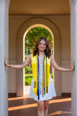 Young woman in white dress with yellow graduation stole and honor cords poses between columns at university.